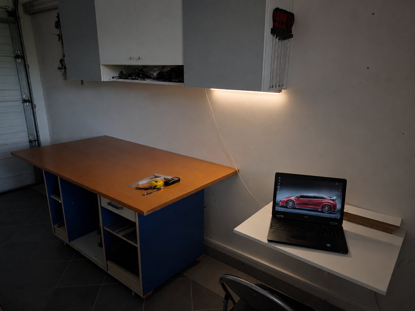 Cleared garage workbench with a tape measure, ruler and utility knife on it, and a laptop set up on a wall shelf beside it